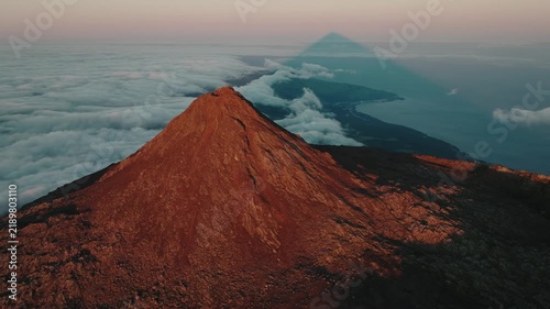Majestic aerial perspective of the Mount Pico volcano summit at sunset, highlighting its perfect triangular shadow cast over the Atlantic cloud layer and the island's coastline.