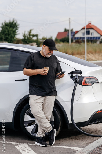 Man near a connected white electric car at an outdoor charging station, illustrating everyday EV use, sustainable transportation and road travel.