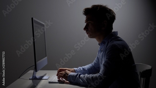 Man with a concentrated expression working on a computer in a dimly lit environment, symbolizing cybersecurity, digital crime, or intense focus on online tasks