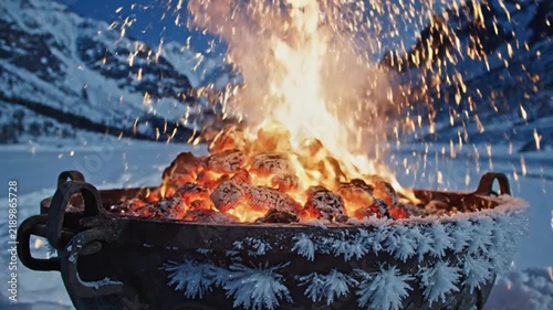 A warm campfire burns brightly in a frozen metal container surrounded by snowy mountains in a cold winter landscape