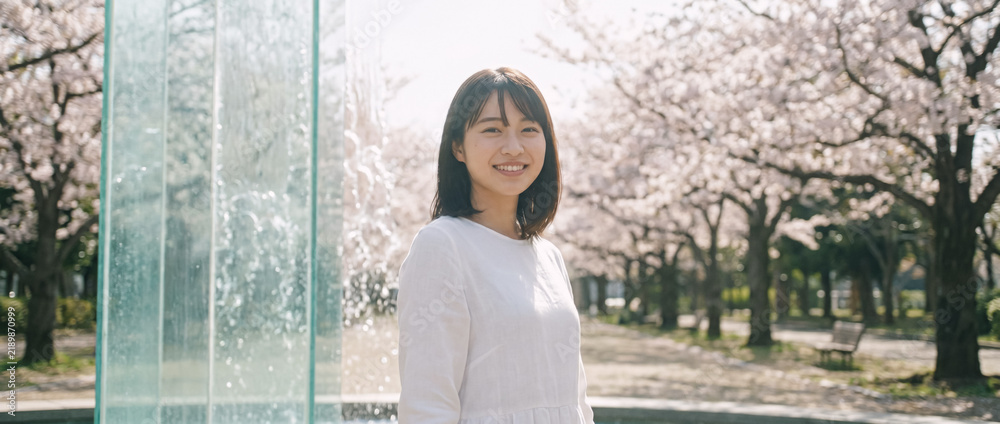 Obraz premium Smiling young Japanese woman by a fountain in a park with cherry blossoms in full bloom