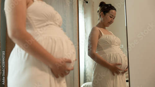 Картината върху платно Close-up of a pregnant woman in a white dress looking down tenderly at her belly, reflected in the mirror