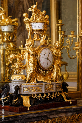 Obraz na plátně Close-up of a highly ornate antique clock featuring gilded bronze figures, cherubs, and marble details inside the Palace of Versailles