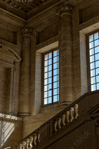 Fotografie Warm sunlight shining through tall windows onto a grand stone staircase and classic columns inside the historic Palace of Versailles, France