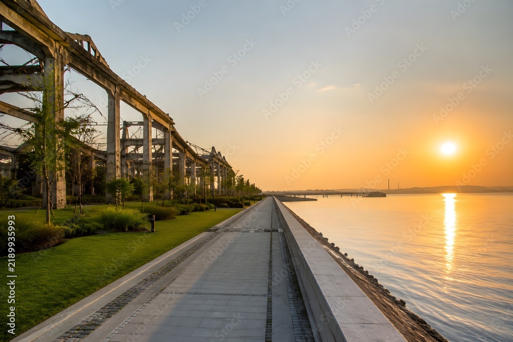 Fototapeta premium Reconstructed waterfront promenade beside damaged warehouse shells converted into green open spaces with calm water and sunrise light