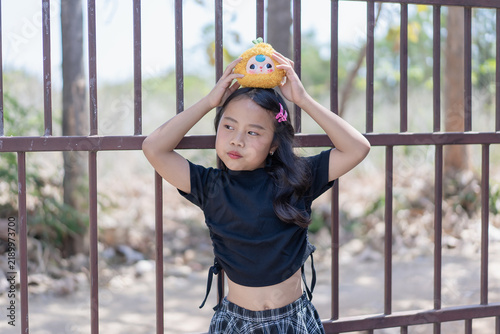 Portrait cute little girl holding plush toy on head black shirt plaid skirt metal gate outdoor playful youth childhood innocent fun lifestyle expression happy kid