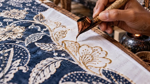 Close up of an artisan hand using a canting tool to apply hot wax on traditional batik fabric.