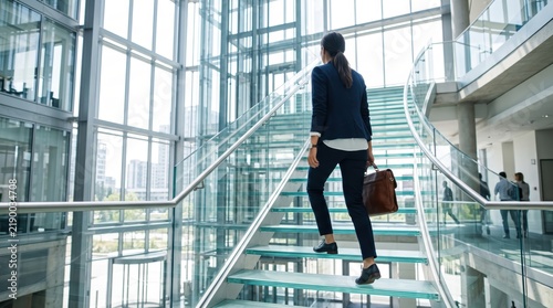Professional businesswoman climbing a modern glass staircase in a bright corporate building lobby, carrying a leather briefcase, symbolizing career advancement and upward mobility.