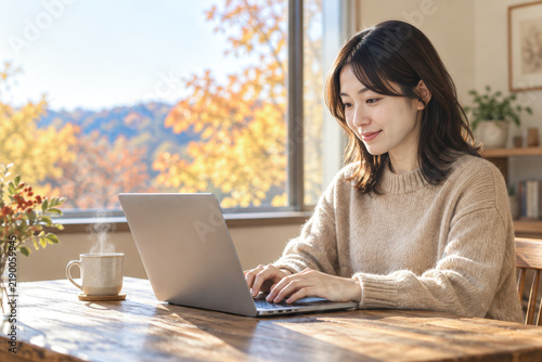 窓外の紅葉を背景にノートパソコンで在宅ワークをする女性