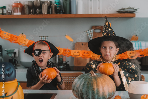 Portrait of a girl and her a little brother celebrating Halloween and decorating pumpkins in the kitchen. They look scary in witch and pirate costumes. Kids Halloween party.