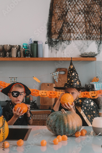 Portrait of a girl and her a little brother celebrating Halloween and decorating pumpkins in the kitchen. They look scary in witch and pirate costumes. Kids Halloween party.