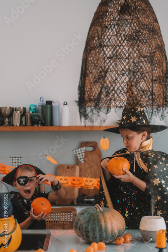 Portrait of a girl and her a little brother celebrating Halloween and decorating pumpkins in the kitchen. They look scary in witch and pirate costumes. Kids Halloween party.