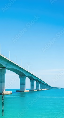 An expansive modern concrete bridge stretches across calm turquoise ocean waters toward the horizon under a clear blue sky at high noon, building, sunny, nature