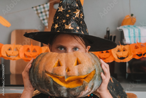 A portrait of a little girl dressed as a witch, holding a large pumpkin with a scary smile carved into it near her head. A children's Halloween party. The main autumn holiday.
