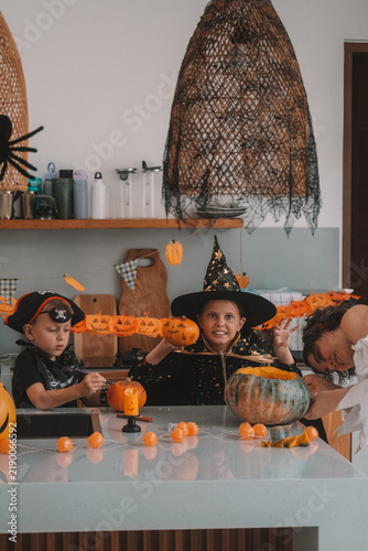 A portrait of a young girl and her younger brother celebrating Halloween and carving pumpkins in the kitchen with their mother's help. They are dressed in witch and pirate costumes. 