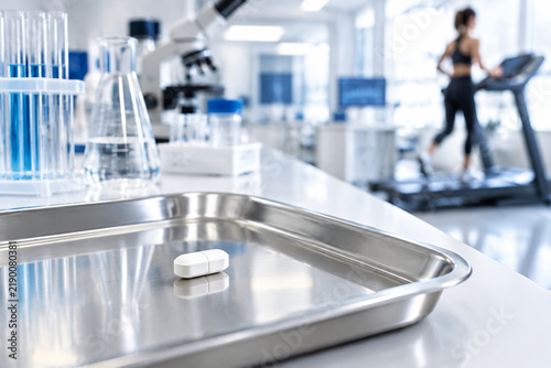 White pill on metal lab tray with exercise equipment in background, medical and fitness concept