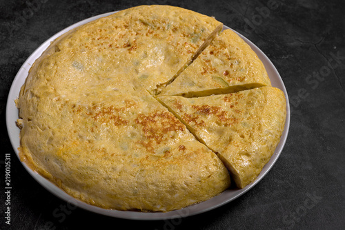 Traditional Spanish omelette served on a plate with slice cut, fresh bread, olive oil, and raw ingredients on a rustic dark surface