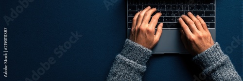 Hands of a person wearing a gray sweater typing on a laptop keyboard against a dark blue background, showcasing a modern workspace setup