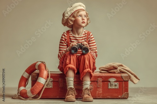 Young girl dressed in vintage sailor attire holding binoculars sits on a red suitcase with a lifebuoy and blanket beside her in a neutral studio setting