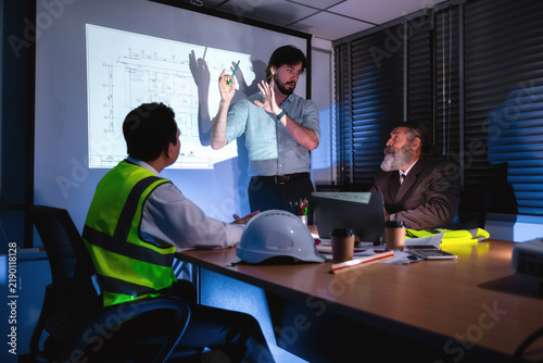 Engineering presenting and discuss floor plans on projector screen during late-night company meeting with senior project manager. Concept of brainstorming in construction project management with team.