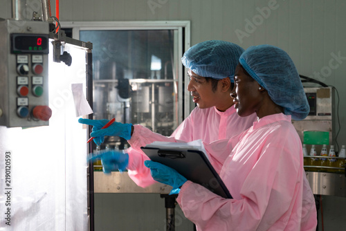 Asian male and black woman factory workers monitoring cold drink bottles on production line at drinks production plant.