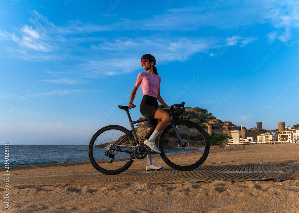 Fototapeta premium Full-length shot of a smiling female cyclist facing the sunrise, standing astride her road bike on a sandy beach, with the blue sky and the medieval town of Tossa de Mar in the background.
