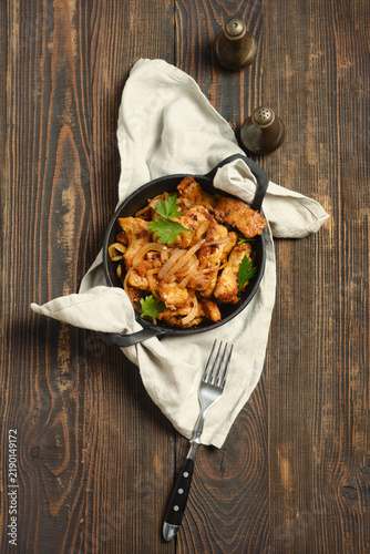 Pork stir fry mixed with onion in hot cast iron skillet placed on wooden table. Fork and napkin are beside pan.