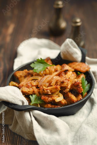 Close-up view of pork stir fry with sliced onions and fresh parsley in cast iron skillet. Dish is served on light cloth placed on wooden table.