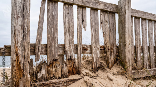 Weathered wooden fence on beach dune eroding, showing old boards and sand accumulating near ocean water