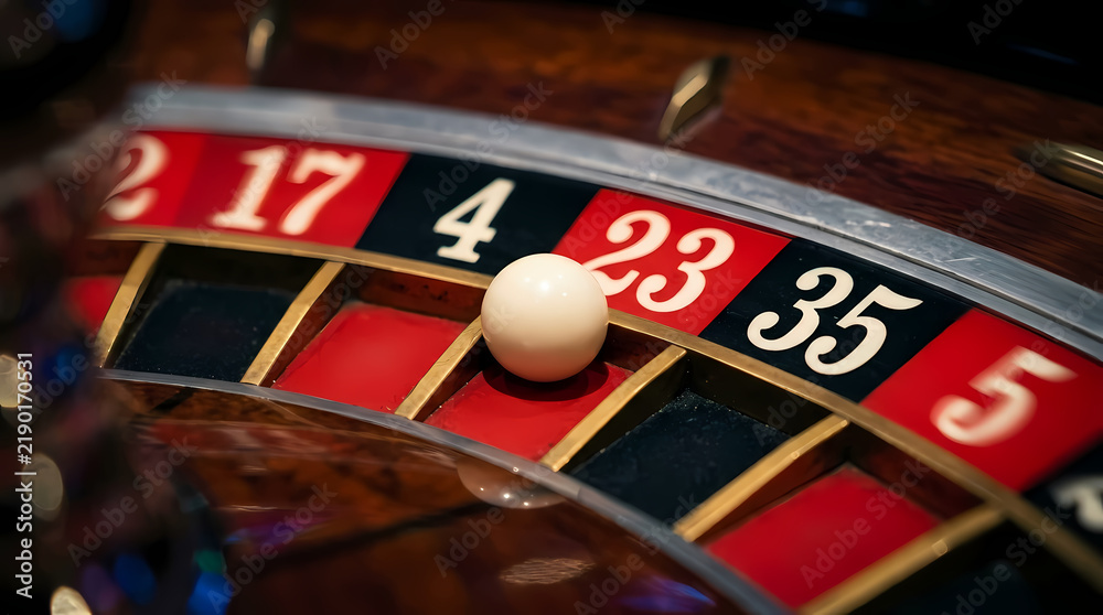Obraz premium A small white ball spins and lands on number twenty three inside a wooden casino wheel while players wait for the outcome of their bets in a tense atmosphere during a night of gaming at a club