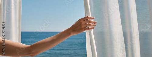 Woman hand open white curtain to see beautiful sea view on a sunny day