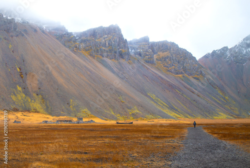 Hiker walking on gravel path towards Viking village film set under steep Vestrahorn mountain in Stokksnes, Iceland