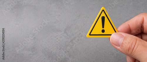 Hand holding yellow triangular warning sign with black exclamation mark on textured gray concrete background, danger alert and caution concept with copy space for text