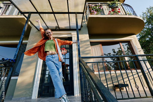 Young woman rejoices outside her apartment building  raising arms happily. She wears casual orange shirt and blue jeans. Concept of joyful modern urban lifestyle.