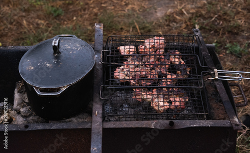 Konstfotografi Juicy meat grilling in a metal wire grate over hot coals with black cast iron pot on BBQ grill outdoors