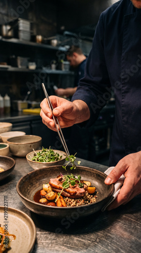 Professional chef placing delicate microgreens with tweezers on gourmet dish in kitchen
