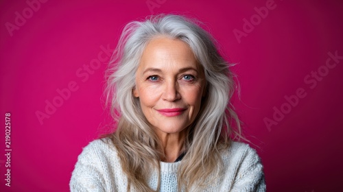 A graceful older woman with flowing silver hair and a captivating smile poses confidently against a vibrant pink backdrop, embodying beauty and self-assuredness.