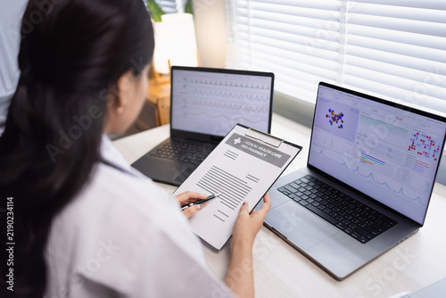 A female doctor working in a hospital office, reviewing medical data, charts, and healthcare reports on dual laptops and a clipboard.