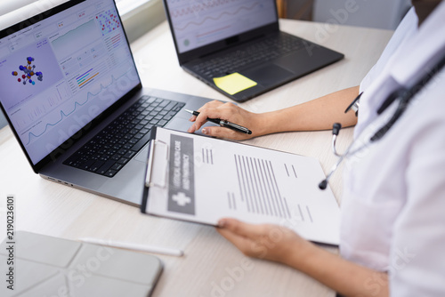 A female doctor working in a hospital office, reviewing medical data, charts, and healthcare reports on dual laptops and a clipboard.