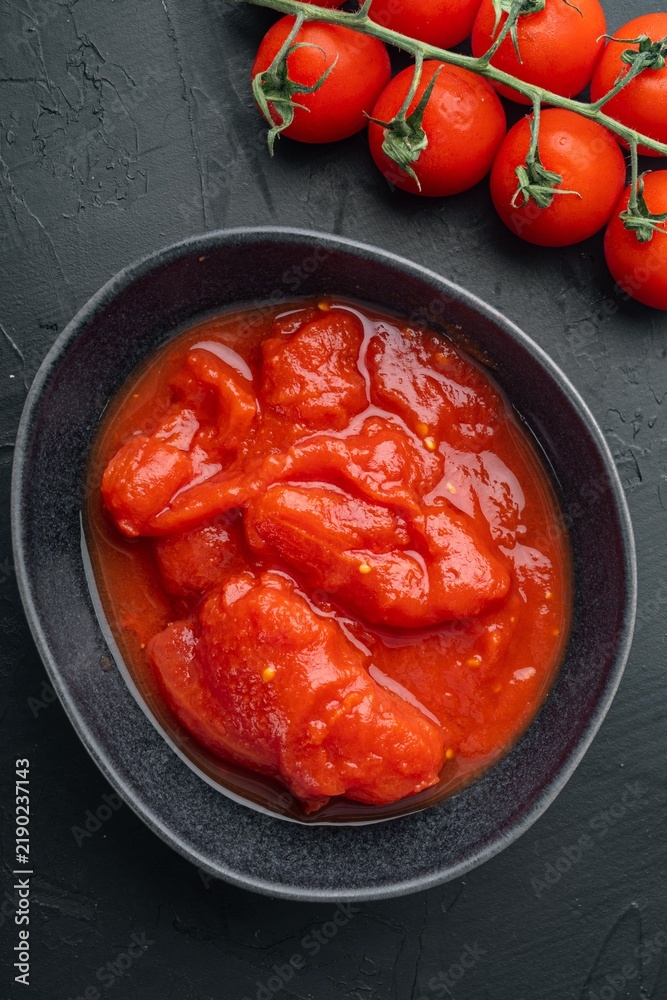 Obraz premium Canned tomato chunks in dark ceramic bowl beside fresh vine tomatoes on black textured surface under soft directional lighting