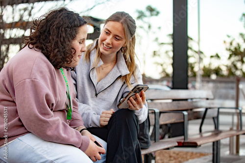Two young women engaged in conversation while sitting on an urban bench looking at a smartphone