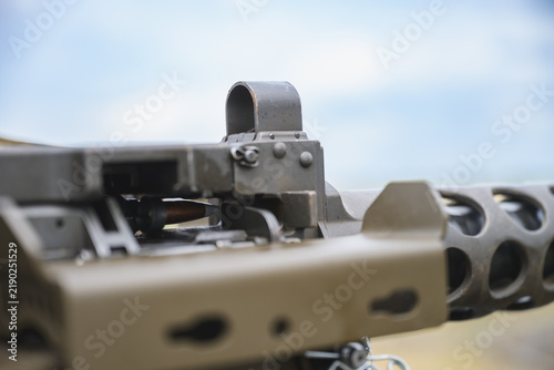 Obraz na plátně Detailed close up of a mounted heavy machine gun showing the metal iron sight, receiver and perforated barrel assembly against a softly blurred outdoor background and blue sky