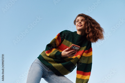 Smiling woman in a colorful striped sweater holds a phone  standing outdoors against a blue sky. Casual fashion  confidence  and carefree energy captured in bright daylight.