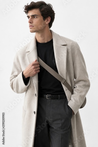 Confident young man in stylish beige trench coat and black shirt adjusting gray belt strap, urban street fashion, minimalist portrait with neutral background, modern casual look