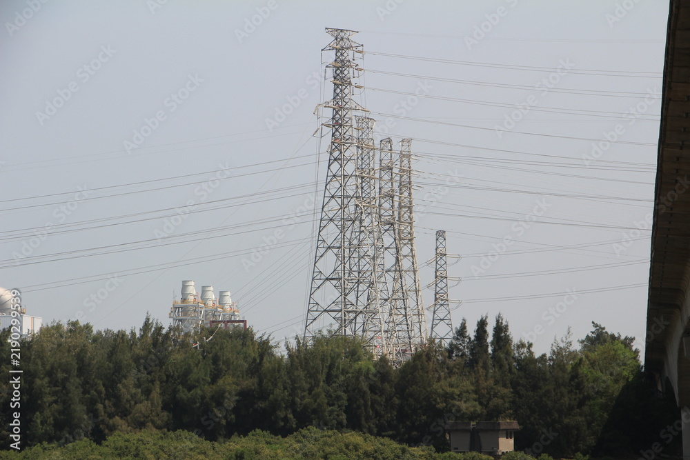 Obraz premium High voltage power transmission towers and pylon electricity lines near industrial power plant complex behind green trees