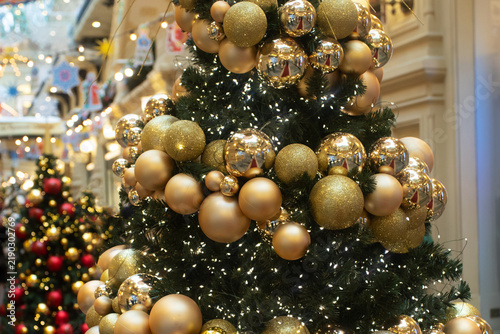 decorated christmas tree with shiny and matte gold baubles, glitter spheres and twinkling lights, festive installation inside a shopping arcade with blurred holiday decorations