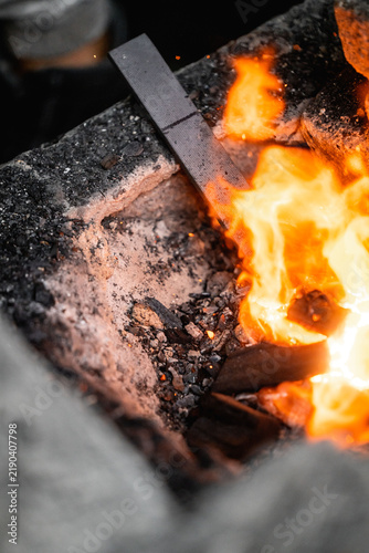 Close-up of a hot metal bar heating in a coal forge with bright orange flames, glowing embers and ash in a blacksmith's workshop