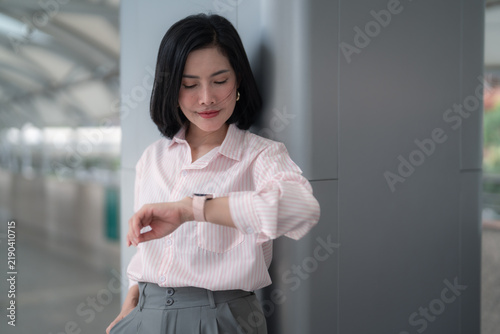 Businesswoman in a pink striped shirt checking the time on her smartwatch while leaning against a wall, waiting and punctuality concept.
