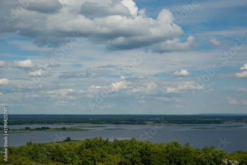 A view of the Dnipro river, with dense green forest on the near shore in the foreground, islands on the water, a distant shore beyond the river, and blue sky with fluffy white clouds above.