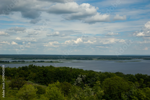 A view of the Dnipro river, with dense green forest on the near shore in the foreground, islands on the water, a distant shore beyond the river, and blue sky with fluffy white clouds above.